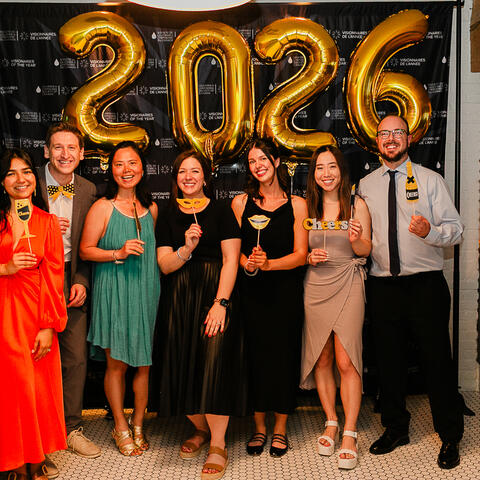 Seven participants pose with playful props beside gold 2026 balloons during the Visionaries celebration.