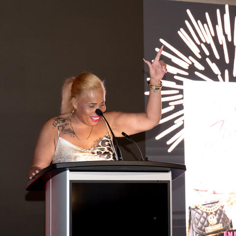 A speaker stands at the podium on stage, raising an arm while addressing guests during the event program.