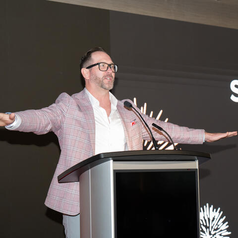A speaker at the podium extends both arms while announcing the silent auction is closing during the evening program.