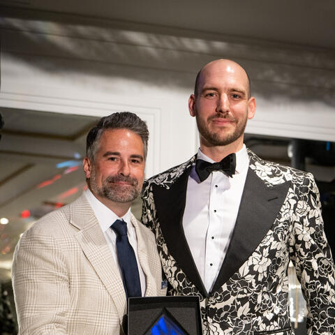 Participant stands holding an award and posing closely during a celebratory moment at a formal fundraising event indoors.