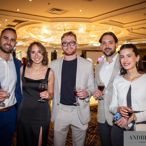 Participants gather in a ballroom holding drinks and smiling together during a lively fundraising event under warm lights.