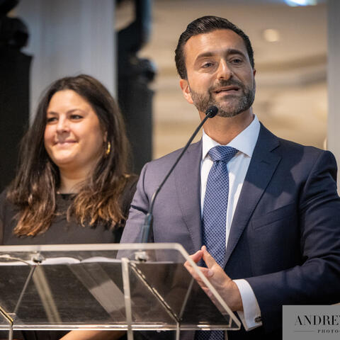 A speaker addresses participants at a podium while another participant stands nearby on stage during a formal fundraising event.
