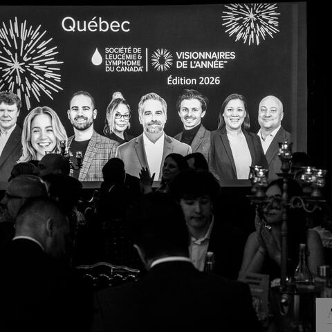 Participants watch and applaud as a large stage screen highlights honorees at a fundraising gala in a dimly lit ballroom.
