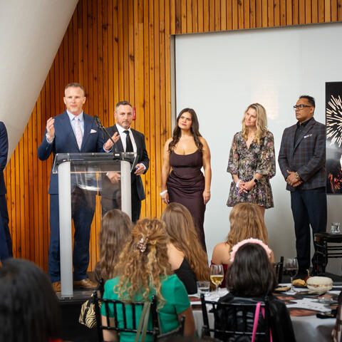 A speaker presents at a podium while participants stand nearby and attendees seated at tables watch attentively.