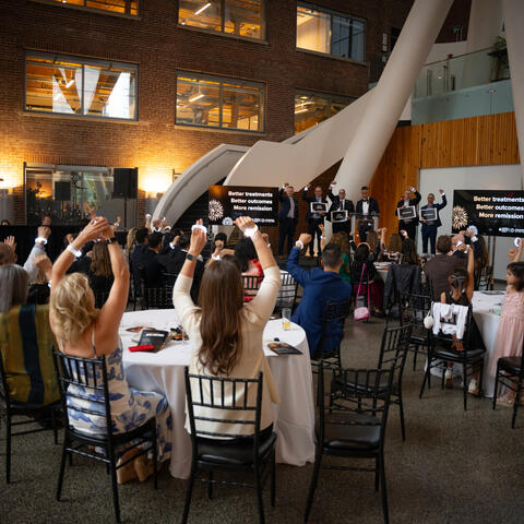 A speaker stands at a podium addressing attendees as a presentation screen displays images during the event program.