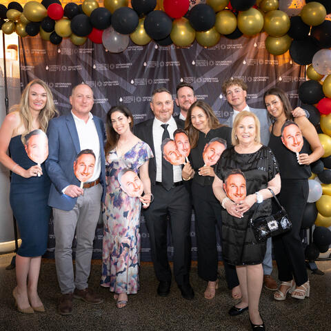 Participants gather closely holding face cutouts while posing together in front of the event backdrop.