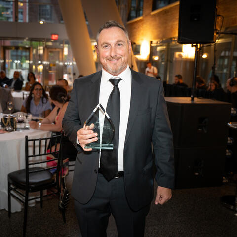 A participant stands in the event space holding an award while attendees seated at tables look on during the program.